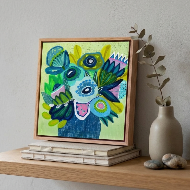 A colorful framed abstract painting featuring a stylized face in shades of blue, green, yellow, and pink, displayed on a light wood shelf next to a beige ceramic vase with dried leaves.