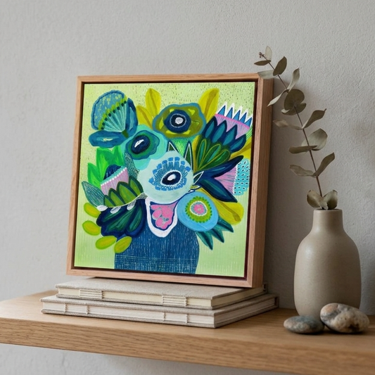 A colorful framed abstract painting featuring a stylized face in shades of blue, green, yellow, and pink, displayed on a light wood shelf next to a beige ceramic vase with dried leaves.