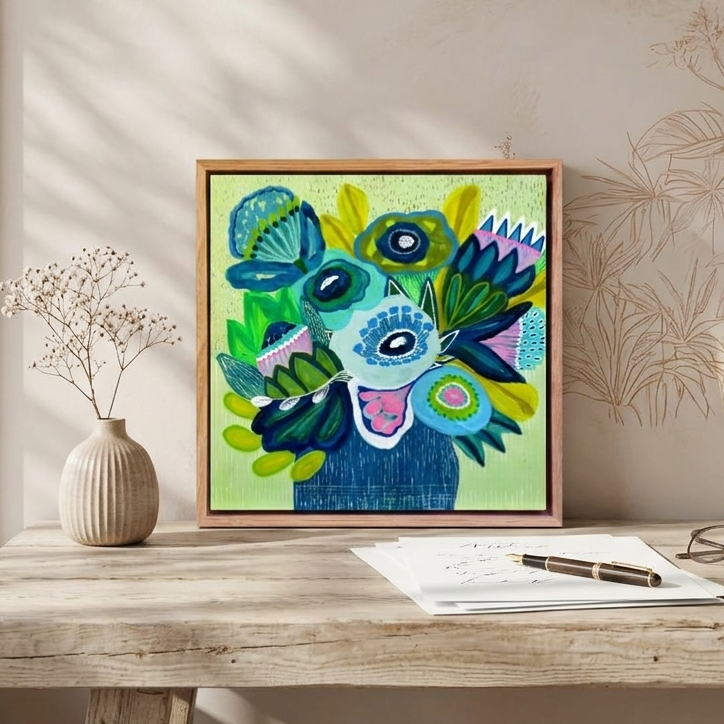 A framed colorful abstract floral artwork sits on a rustic wooden table in a styled room, with a vase, glasses, and papers nearby. The artwork features blue, green, purple, and yellow abstract shapes on a light green background.