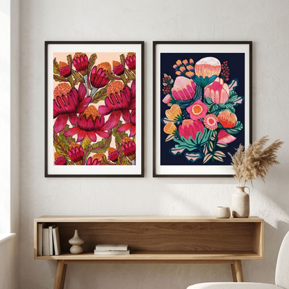 Australian Bush Blooms wall art print set by Kirsten Katz: two framed botanical prints, red waratahs on soft blush and a protea and banksia bouquet on midnight navy, hung above a timber console
