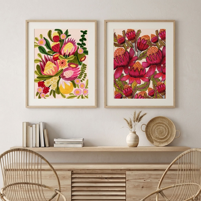 Waratahs & Proteas wall art print set by Kirsten Katz: two framed botanical prints of Australian proteas, native flowers and red waratahs on soft blush grounds, hung above a timber shelf with rattan chairs