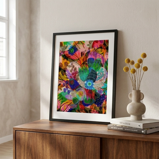 Colorful modern abstract art print in a black frame on a wooden surface with a vase and books.