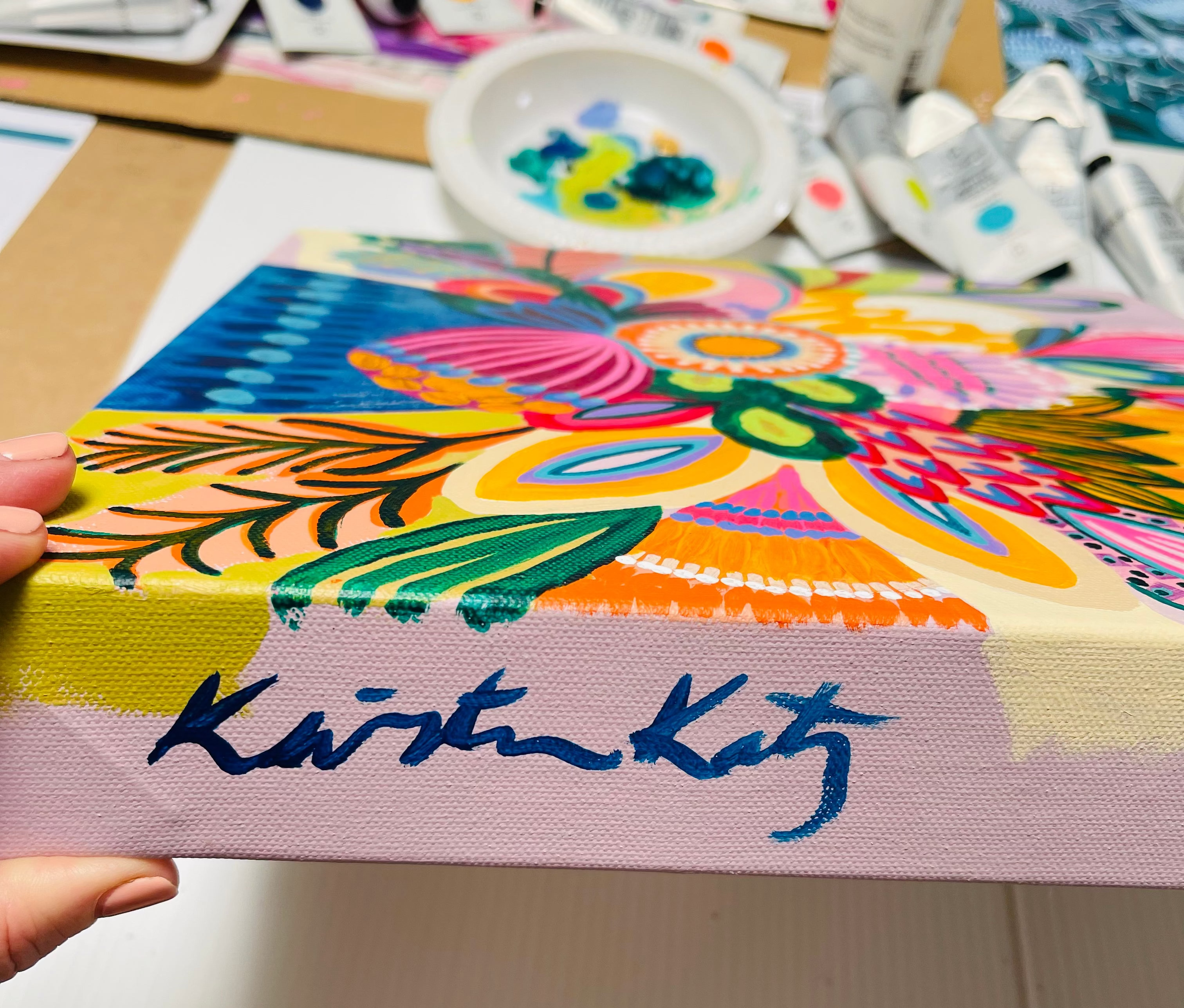 Colorful artwork with 'Kirstin Katz' signature held by a hand on a blurred background.